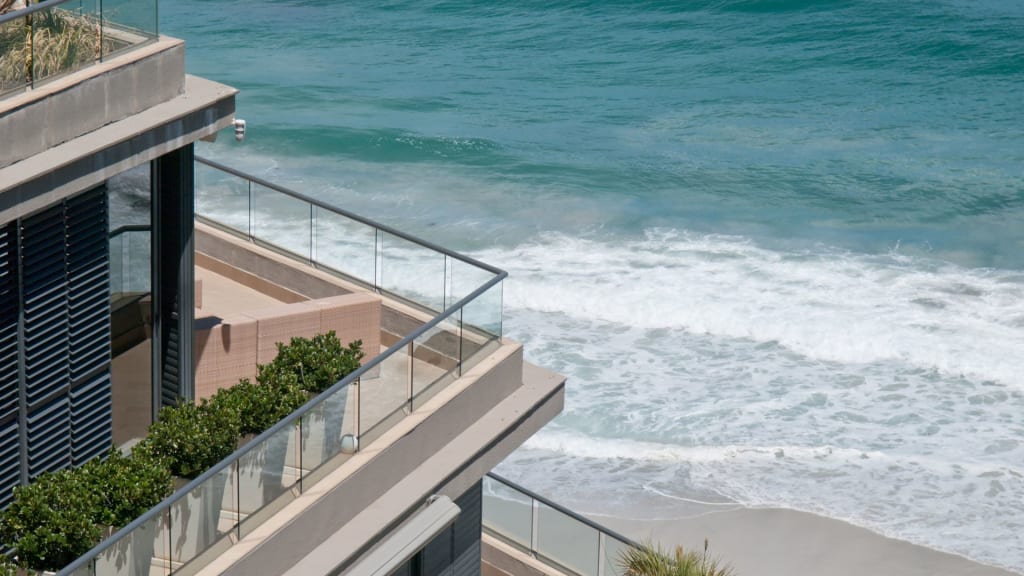 Modern beachfront building with glass balconies overlooks ocean waves gently crashing onto a sandy shore under daylight.
